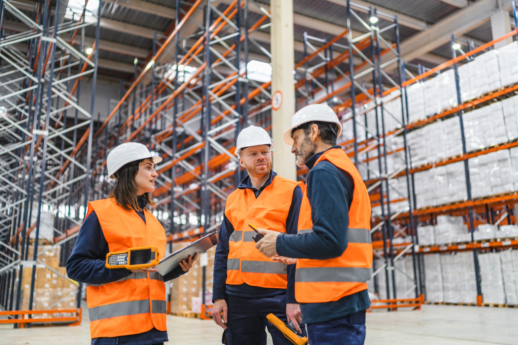 Three factory workers in orange vests and hard hats stand in a factory talking to each other.