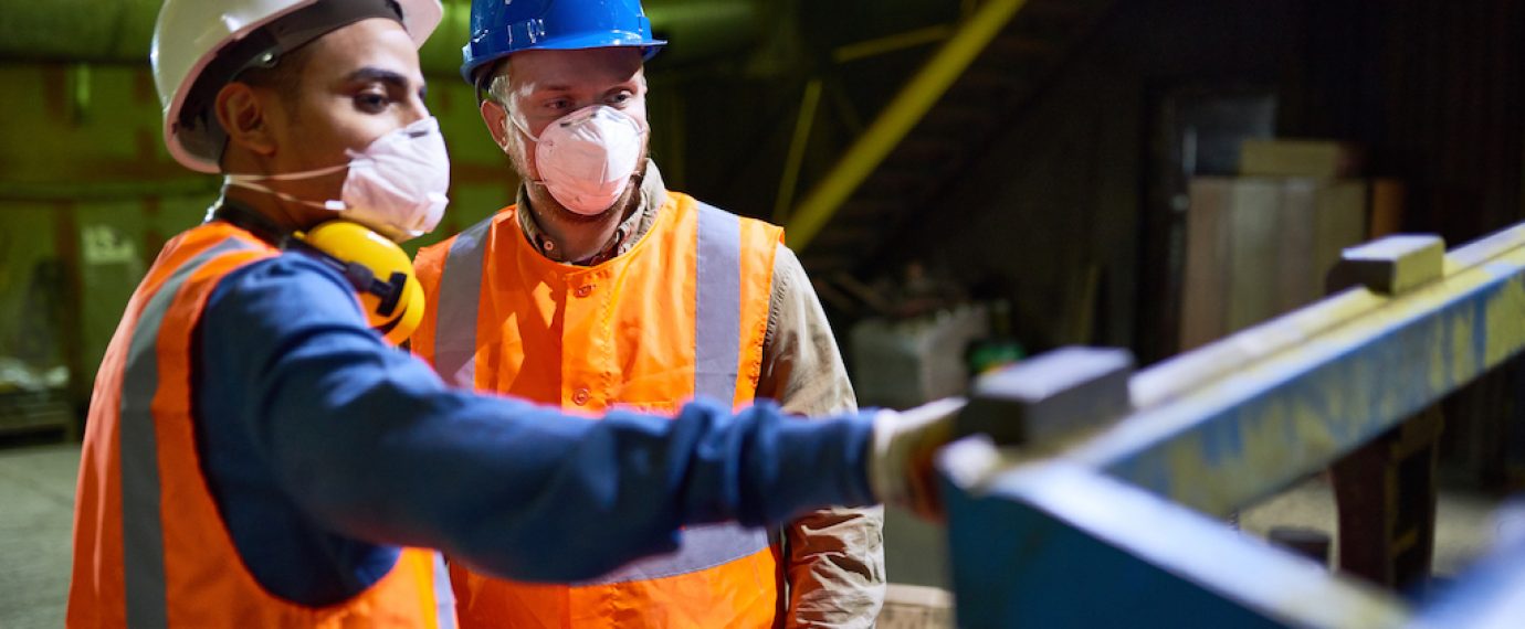 Handsome mixed race technician wearing reflective vest pointing at something while discussing results of joint work with his colleague, interior of spacious production department on background