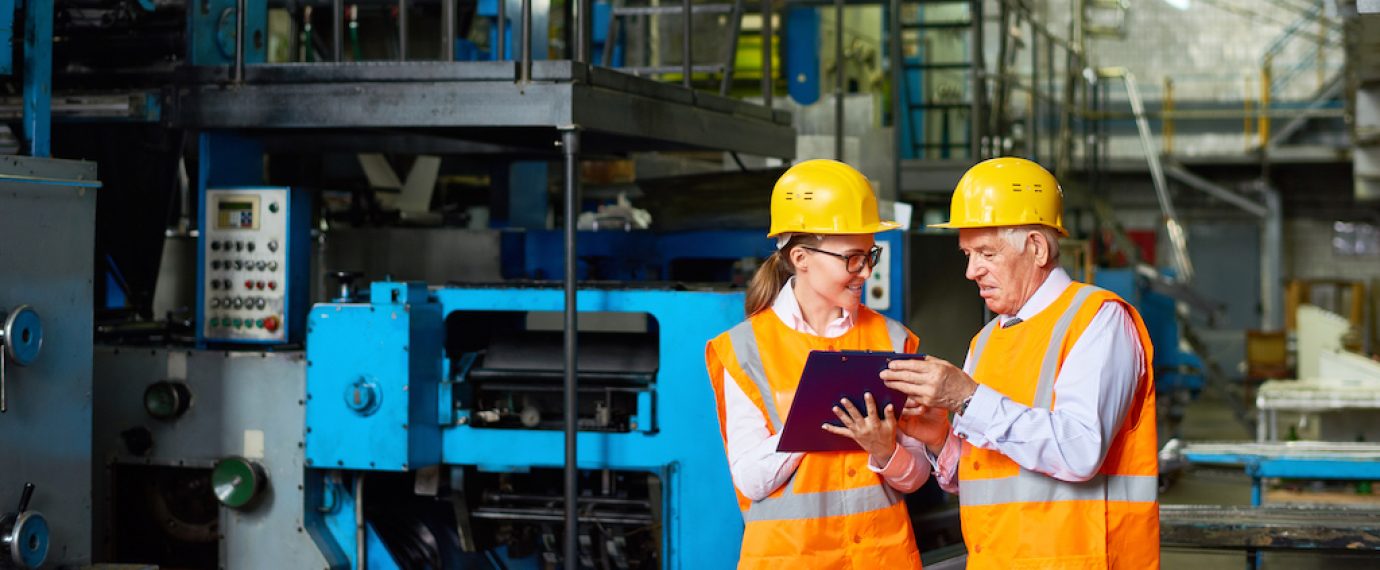 Portrait o senior factory worker discussing something with young woman in  modern workshop,  both wearing hardhats and reflective jackets