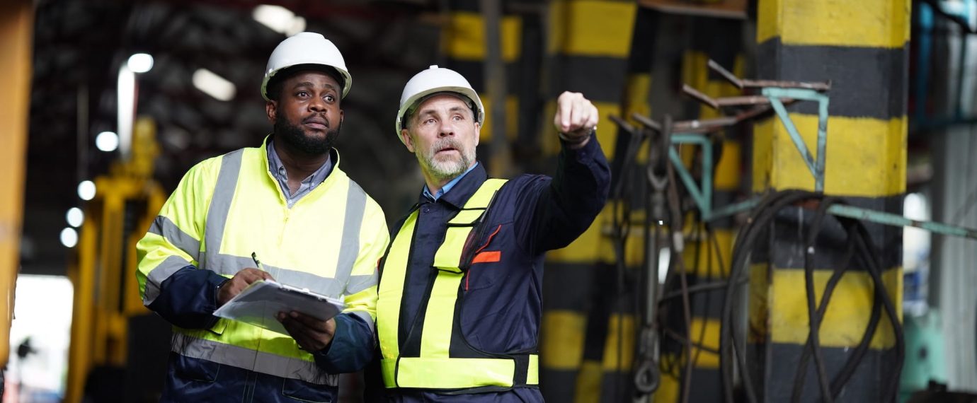 Two factory workers in safety vests and hard hats pointing and looking