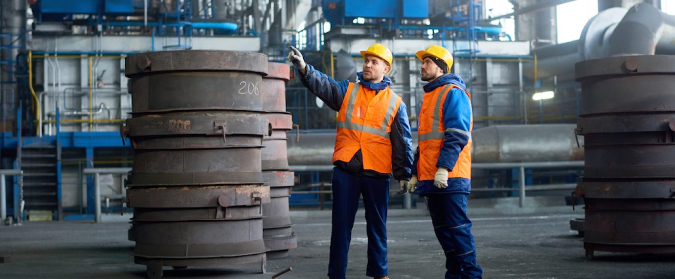 Bearded inspector wearing reflective vest and hardhat pointing at something while young technician giving tour of modern factory, interior of spacious production department on background
