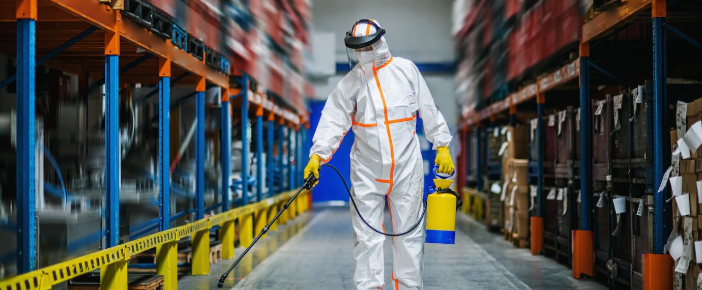 Man worker with protective mask and suit disinfecting industrial factory with spray gun.